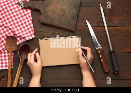 open notebook with blank brown sheets and kitchen utensils on a brown wooden table, female hands hold a black pencil, top view Stock Photo