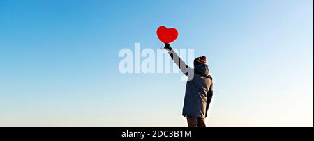 young man from the back in warm clothes holding a red heart-shaped balloon against the blue sky. Valentine's day and romantic concept, copy space Stock Photo