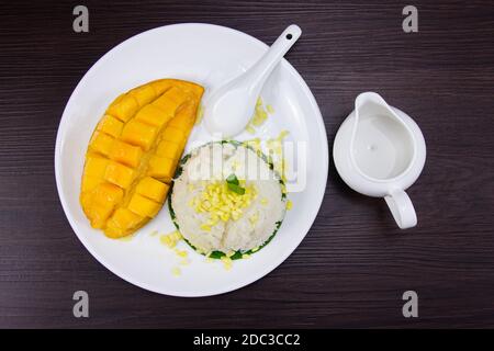 Sticky rice with fresh ripe mango with coconut milk put white plate on brown dark wooden table. Top view. Stock Photo