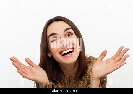 Excited young woman , casual in a golden sweater, screams happily, raises her hands up in surprise, over white studio wall with copy space.  Stock Photo