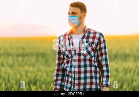 A young man in a checked shirt, with a medical protective mask on his face, stands in a wheat field against the setting sun Stock Photo