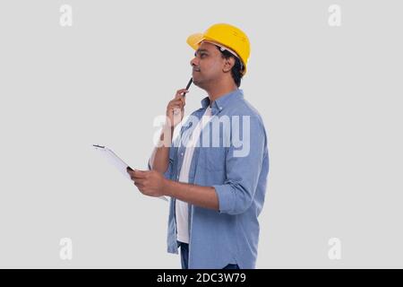 Construction Worker Holding Clipboard in Hands Watching up. Man Thinking. Architect Holding Blueprints. Yellow Hard Helmet. Stock Photo