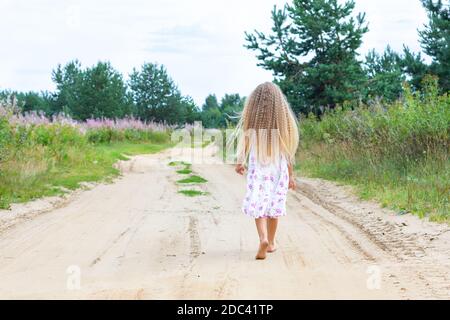 A girl with long blond curly hair walks along a rural road. Full-length portrait from the back. A walk in the fresh air in solitude. Stock Photo