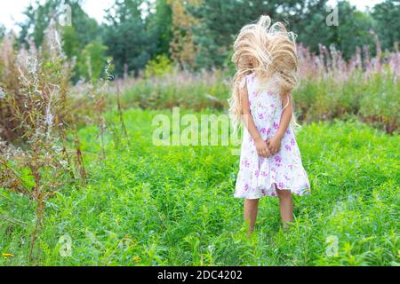 A girl in a white sundress, with lush blond hair, stands in a flowering meadow, shook her head, no face is visible, her hands are folded. Stock Photo