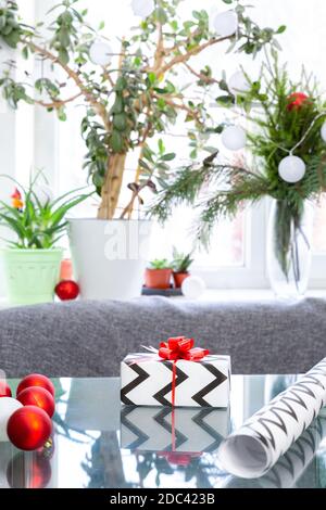 Christmas gift, wrapping paper, Christmas balls on glass table against the background of a window with houseplants and a garland.New Year, Xmas, winte Stock Photo