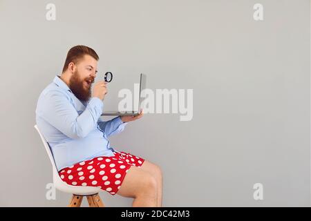 Funny man in red shorts with a laptop increases magnifying information on a gray background. Concept of online freelancer working remotely. Stock Photo