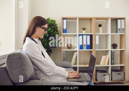 Smiling woman doctor sitting in clinic with laptop on knees and communicating during online consultation Stock Photo