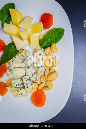 Gourmet snacks in flat lay.Delicious dried apricots,almonds & hazelnuts,brie & blue cheese served on white plate,shot directly from above on black tab Stock Photo