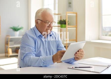 Senior man with glasses sitting at a table at home using a tablet reads information on the Internet. Stock Photo