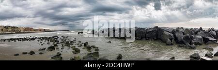 Dramatic weather panorama with cloudy sky and sunbeams between clouds, low angle view in a rocky bay with city skyline. Stock Photo