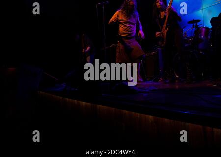 Male vocalist dancing wearing kilt while guitarist, bassist and drummer perform passionately illuminated with blue light Stock Photo