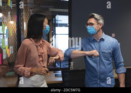 Caucasian man and asian woman wearing face masks greeting each other by touching elbows at modern of