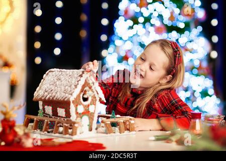 Kids baking Christmas gingerbread house. Children celebrating winter holiday at home. Decorated living room with fireplace and tree. Family activity. Stock Photo