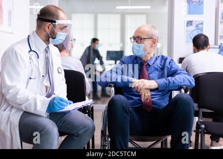 Doctor wearing face mask and visor against infection with coronavirus while talking with disabled senior man in waiting area sitting on wheelchair. Patient and nurse at clinic reception. Stock Photo