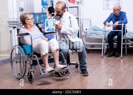 Doctor showing brain X ray image of disabled elderly senior woman in wheelchair. Man with disabilities ,walking frame sitting in hospital bed. Health care system, clinic patients. Stock Photo