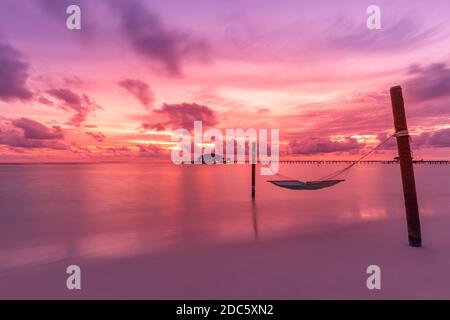 Dramatic sunset sky in tropical island beach. Swing or hammock over water. Purple pink sky clouds, artistic sunrise landscape, coastline Stock Photo