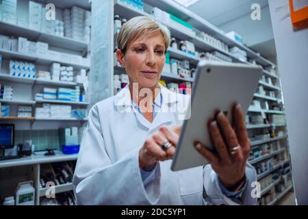 Senior female pharmacist scrolling through prescriptions on digital tablet standing in pharmacy  Stock Photo