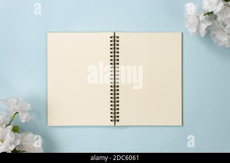 Top Table Spiral Notebook or Spring Notebook Mockup in Unlined Type on Center Frame and White Flowers at Bottom Left and Top Right Corner on Blue Past Stock Photo