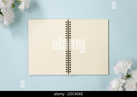 Top Table Spiral Notebook or Spring Notebook Mockup in Unlined Type on Center Frame and White Flowers at Bottom Right and Top Left Corner on Blue Past Stock Photo