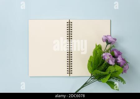 Top Table Spiral Notebook or Spring Notebook Mockup in Unlined Type on Center Frame and Purple Flowers at Bottom Right Corner on Blue Pastel Minimalis Stock Photo