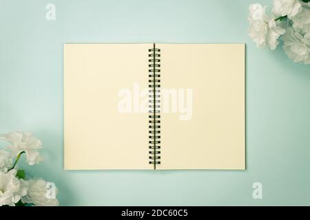 Top Table Spiral Notebook or Spring Notebook Mockup in Unlined Type on Center Frame and White Flowers at Bottom Left and Top Right Corner on Blue Past Stock Photo