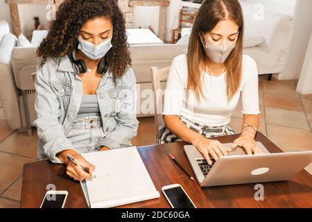 Two young mixed race beautiful women working from home sitting on a wooden table in elegant living room wearing Coronavirus safe protection mask - Col Stock Photo