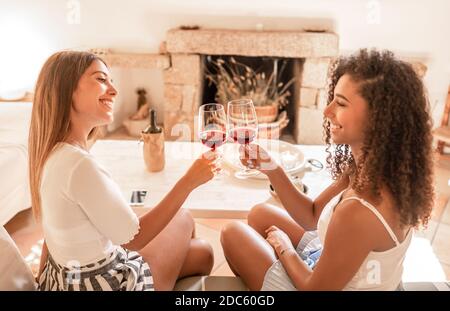Two young mixed race beautiful women toasting at home with red wine glasses - Smiling female couple of friends sitting on sofa looking each other in e Stock Photo