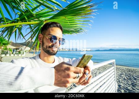 Handsome Caucasian bearded man with round sunglasses using smartphone under a palm on the beach - Stylish male person texting on phone messaging his f Stock Photo