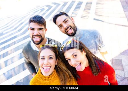 Two Caucasian couple looking at the camera from the bottom smiling and making mouths open faces sticking out their tongues - Friends having fun outdoo Stock Photo