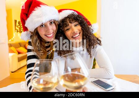 Two young beautiful mixed race women wearing Santa hat looking at the camera holding white wine glasses - Female lgbt couple smiling toasting with cha Stock Photo