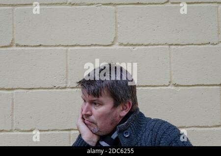 Portrait of man who put his chin on his palm outside. Middle-aged grayish man sitting in thoughtful pose against yellow wall background. Stock Photo
