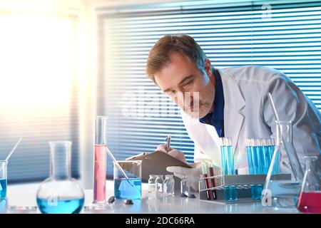 Scientist writing in a notebook supported on a laboratory with glassware and windows background. Horizontal composition. Front view.