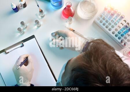 Laboratory technician writing on notepad  with vial in hand on a laboratory table. Top view. Horizontal composition. Stock Photo