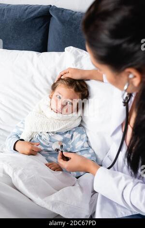 doctor touching head of diseased girl, lying in bed, and examining her with stethoscope Stock Photo