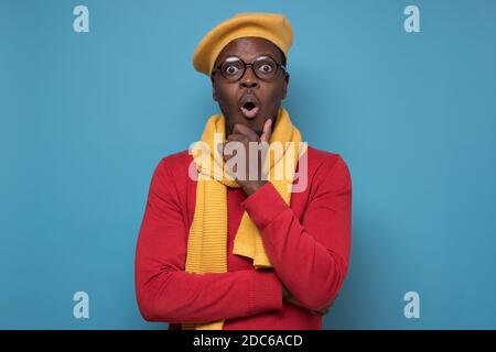 Young African American man wearing glasses and hat looking surprised. Studio shot on blue wall. Stock Photo