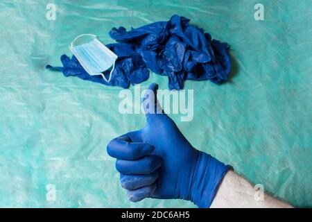 A hand with thumb up as an approval gesture with an unfocused pile of personal care equipment worn out in healthcare placed on green table. Stock Photo