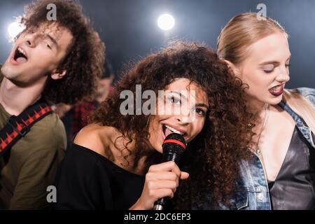 Happy rock band vocalist with microphone singing near musicians with backlit on blurred background Stock Photo
