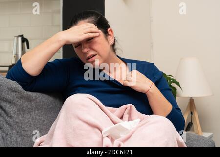 Adult female model feeling sick at home having high body temperature or fever laying on sofa covered with blanket as covid19 influneza symptoms Stock Photo