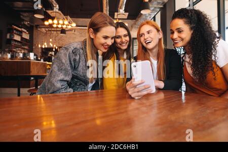 Multiracial female friends having fun at cafe. Group of young women sitting at restaurant and looking at a mobile phone. Stock Photo