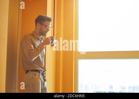 Resting at work. Young handsome businessman drinking coffee or tea while standing in the office and looking out the window Stock Photo