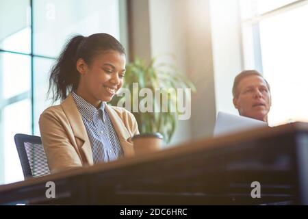 Female office worker. Young happy mixed race woman using laptop and smiling while sitting with male colleague at desk in the modern office Stock Photo