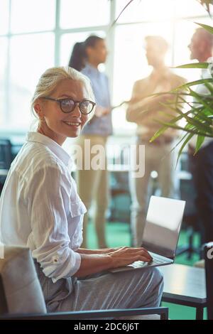 Young cheerful mature business woman sitting in the modern office and using laptop, smiling at camera while her colleagues talking on the background Stock Photo