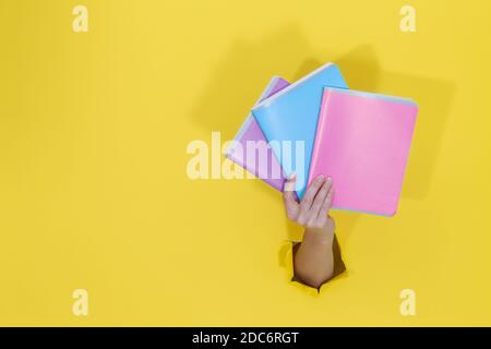 Female hand holding colorful notebooks from yellow torn paper. Back to school Education concept. School stationery and office supplies. Space for text Stock Photo