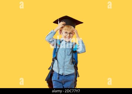 Child boy primary school student in a graduate cap, glasses and with a backpack on a yellow background. Knowledge day and back to school. Copy space Stock Photo
