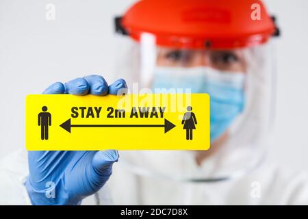 Medical worker in personal protective equipment holding yellow 'STAY 2m AWAY' sign,social physical distancing to prevent and stop spread of viruses