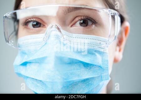 Closeup of a worried female NHS UK doctor or lab scientist wearing PPE safety glasses & blue face mask,COVID-19 corona virus disease global pandemic Stock Photo