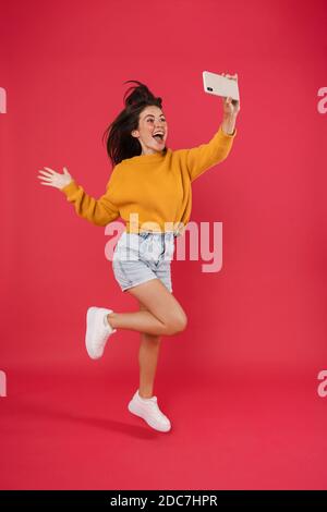 Image of excited brunette girl taking selfie photo on cellphone while jumping isolated over pink background Stock Photo