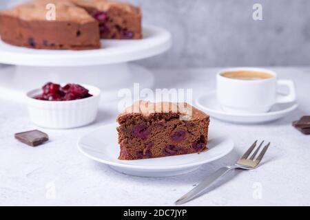 Chocolate cherry pie on a white plate. In the background is a pie, a cup of coffee, cherries and pieces of chocolate. Stock Photo