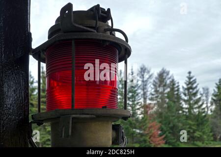A red cylindrical fluted metal-rimmed lantern hanging from a brown wooden post against a blue cloudy sky and green blurred conifers Stock Photo
