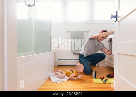 Man fixing sink while daughter lying on hardwood floor in kitchen Stock Photo